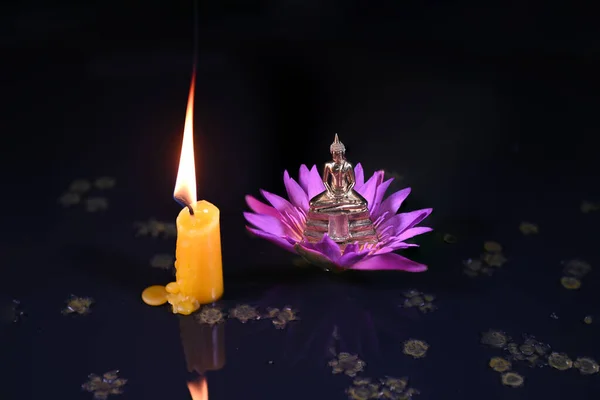 Close-up of a little Buddha statue, water lily, and burning candles on black background