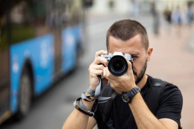 Portrait of young adult caucasian photographer taking a picture with a retro style camera against blurred city street at background
