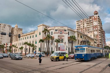 Alexandria 'da bir kadın karşıdan karşıya geçiyor. Siyah ve sarı taksiler, arabalar ve mavi tramvay İskenderiye, Mısır - Mart 2023