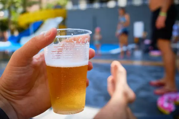 Man holds plastic cup of beer chilling by swimming pool. Hand holds beer at poolside. Summer relaxation