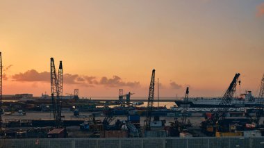 Günbatımında endüstriyel limanın geniş açılı görüntüsü, birden fazla inşaat vinci, kargo konteynırı ve kenetlenmiş gemilerin yer aldığı. Deniz lojistiği ve büyük ölçekli altyapı.