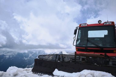 Kırmızı kar temizleme aracı karlı dağ sırtında, dramatik bulutlu gökyüzünün altında duruyor. Karla kaplı tepeleri olan manzaralı alp arka planına yakalanan kar kedisi