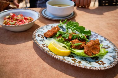 Cig kofte adı verilen geleneksel Türk yemeğinin yakından görüntüsü taze maydanoz ve limon dilimleriyle süslenmiş kıvırcık marul yatağında servis ediliyor. Arka planda domates salatası ve mercimek çorbası var.