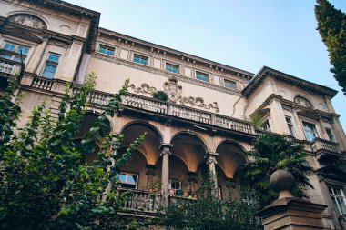 Yeşillik ve klasik mimari unsurların çerçevelediği tarihi malikanenin ayrıntılı cephesi. Simetrik yapıda kemerli pencereler, süslü balkon korkulukları, sütunlar ve heykel kabartmaları yer alıyor