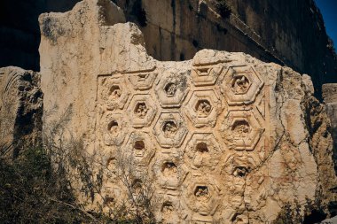 Geometrik çiçek oymalarıyla süslenmiş antik taş duvarın detaylı görüntüsü. Yıpranmış yüzeyde altıgen şekillerin simetrik deseni var. Bu da klasik Greko-Romen mimari etkisini gösteriyor.