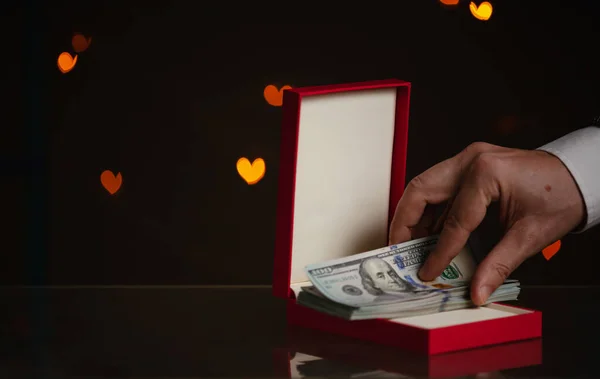 Valentine\'s Day gift. A hand puts a bundle of dollars in a red box. Yellow hearts are glowing behind on a dark backgroun
