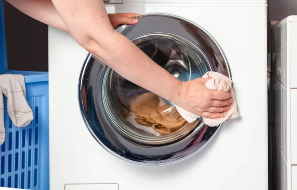 A woman hand wipes the transparent cover of the round hatch of a modern front-loading washing machine with a rag. Laundry is inside the machin