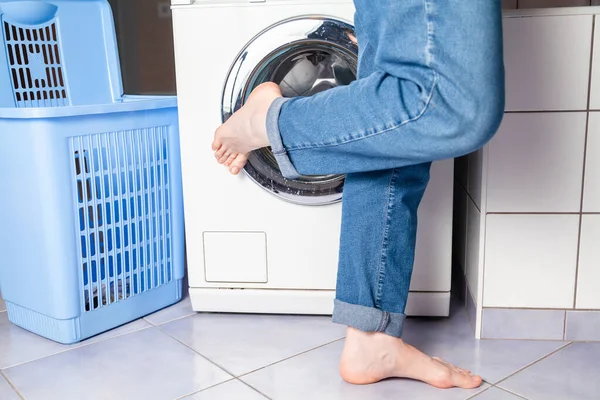 Barefoot female foot closes the transparent lid of a front-loading washing machine. Loaded laundry in the machine. There is a laundry basket nearb
