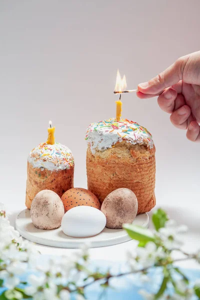 Two Easter cakes and eggs stand on a white background. Candles are inserted into the Easter cakes. A hand lights a candle with a match. Below is a blue cloth and a tree branch with white flower