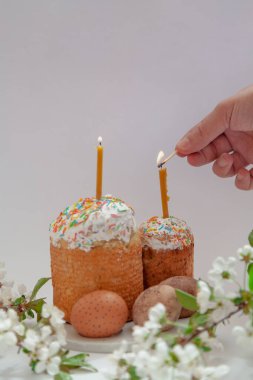 Beyaz arka planda iki Paskalya pastası ve yumurta standı. Mumlar Paskalya pastalarının içine konur. Bir el kibritle bir mum yakar. Ön planda beyaz çiçekli bir ağaç dalı var.