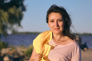 Geniş bir nehrin kumlu sahilinde uzun kızıl saçlı ve parlak sarı ceketli güzel bir kızın portresi.