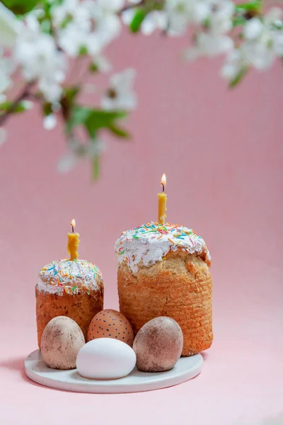 Easter still life. Two Easter cakes and eggs stand on a pink background. Burning candles are inserted into the cakes. Top view of a tree branch with white flower