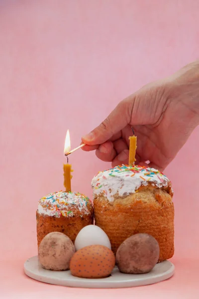 Two Easter cakes and eggs stand on a pink background. Candles are inserted into the Easter cakes. A hand lights a candle with a match. In the foreground is a tree branch with white flower