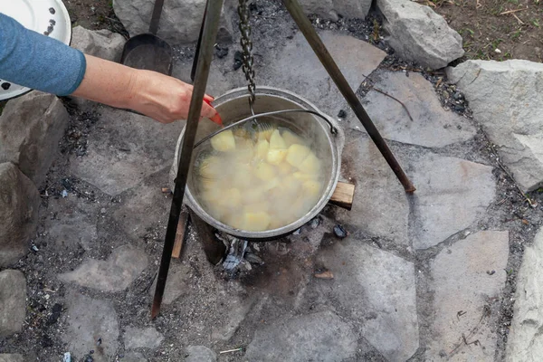 Cooking food over a fire. Hand stirring potatoes in a round tourist pot on a high metal tripod on a stone platfor