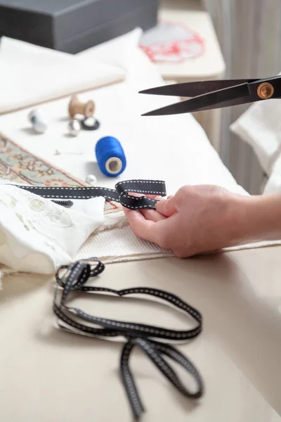 The seamstress hand holds a narrow black ribbon with two white stitches. Large scissors cut off a piece from it. Threads and thimbles lie on the fabri