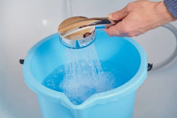 Hand Holds Shiny Shower Head Pours Water Blue Plastic Bucket lizenzfreie Stockfotos