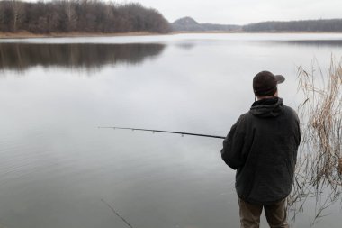 A man in a jacket catches fish with a spinning rod on the shore of a reservoir on an autumn da