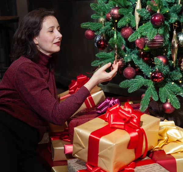 A contented young woman sits in front of a Christmas tree. There are a lot of boxes with gifts in festive packaging on the floor