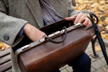 Gri ceketli adam ahşap bir bankta oturuyor. Eskimiş bir stilde kahverengi deri bir çanta açar.