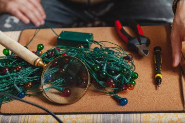 Noel çelengi ve alet. Masanın üzerinde yuvarlak ampullü bükülmüş bir çelenk var. Yakınlarda bir tornavida ve cımbız var. Üzerinde büyük bir büyüteç var.