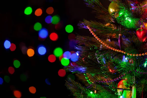 A green Christmas tree festively decorated with toys and luminous garlands stands in a dark room close-up. In the background, colorful bokeh light