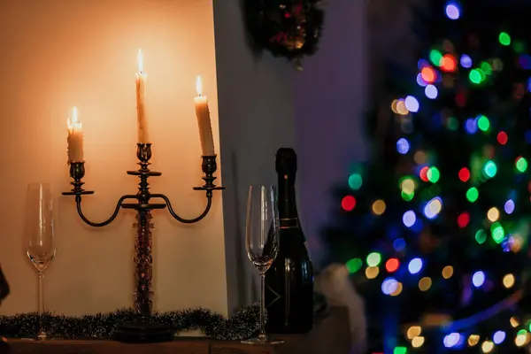 stock image We celebrate the new year at home! A candlestick with three burning candles stands in front of a white wall in a dark room. Nearby is a bottle of champagne and a tall glass. Behind blurry lights on a christmas tre