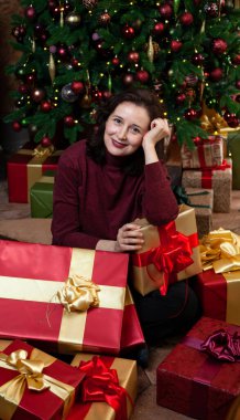 Mutlu genç bir kadın, yeni yıl hediyelerinin önünde tatil paketinde oturur. Arka taraftaki dekore edilmiş Noel ağacı