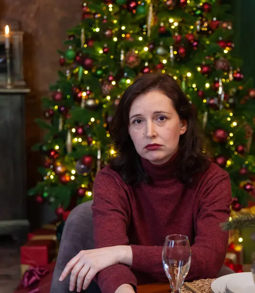 Sad Christmas. Sad young woman sitting alone at a festive table near a decorated Christmas tre
