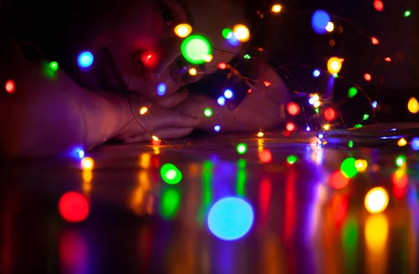 Multi-colored lights of the New Year garland reflect from the smooth surface of the table. In the background in the dark, a woman lies on her folded arms and looks at the light