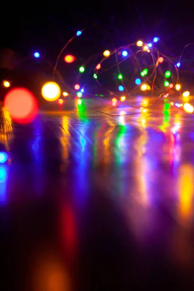 The colorful lights of an electric Christmas garland cast long, shiny paths on a smooth surface. Vertical image on a dark backgroun