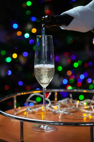 New Year party. Stream of champagne pours into elegant glass from black bottle wrapped in white napkin against background of multi-colored lights of Christmas tree. Bubbles and foam in the glas