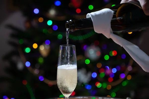 New Year party. A stream of champagne pours into glass from black bottle wrapped in white napkin against background of multi-colored lights of Christmas tree. Bubbles and foam in the glas