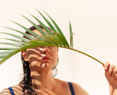 Güzel, büyük boy bir model önünde zarif bir yeşil palmiye dalı tutuyor. Palmiye ağacı kadın gözünü gizler.