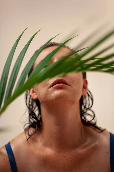 A palm tree branch hides the eyes of a pretty woman in a swimsuit. Long wet hair falls on damp shoulder