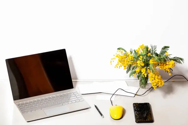 Spring on the computer desk. A yellow computer mouse and a bouquet of yellow mimosa stands on a gray table. There is an open laptop on the left. On the right is a mobile phon