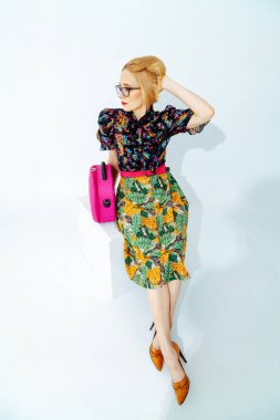 A stylish blonde woman with a short haircut in a skirt with a green floral print sits on a white background. Nearby is a bright pink ba
