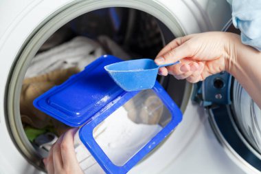 Female hands are picking up washing powder from a blue container with a special measuring container in front of a washing machin