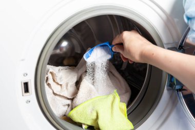 Women hands pour washing powder into the open hatch of a washing machine using a measuring container. Close-u