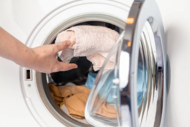 A woman hand throws a dirty towel into the hatch of a modern front-loading washing machin