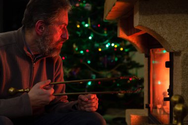 New Year Eve in front of the fireplace. The man looks at the fire in the burning fireplace. In the background, New Year lights are lit on the tree. He holds a fireplace tongs in his han clipart