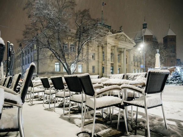 View of a restaurant with chairs and table covered in snow on a cold winter night with magnificent Haussmannian building palace in background
