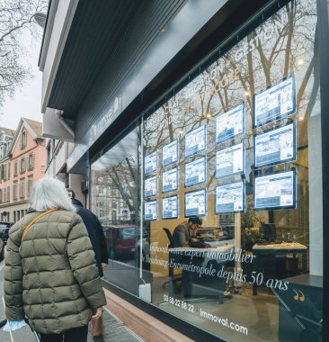 Strasbourg, France - Jan 5 2023: Senior woman looking at the prices of houses and apartments at a real estate agency glass showcase window with multiple announcement