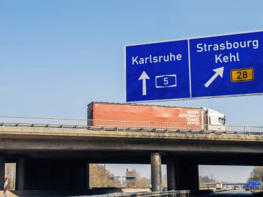 Germany - Feb 10, 2023: Karlsruhe, Strasbourg and Kehl city names on blue highway sign with truck on a bridge - low angle view from the autobahn