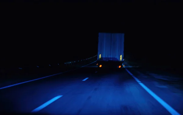 A truck drives along a dark highway, illuminated only by its headlight in the night cityscape.