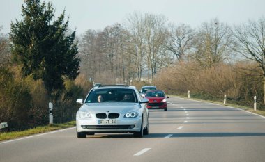 Almanya - 10 Şubat 2023: Bir otoyol boyunca ilerleyen gümüş renkli BMW, ağaçların ve kırsal bitkilerin manzarasını sunuyor. Şehirde etkili bir ulaşım aracı..