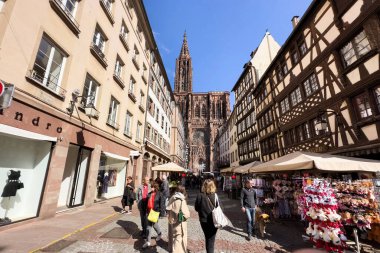 Strasbourg, Fransa - 21 Eylül 2022: Görkemli bir Notre-Dame de Strasbourg katedrali yayaların gezdiği ve hediyelik eşya dükkanlarını ziyaret ettiği kalabalık bir caddeye hakim..