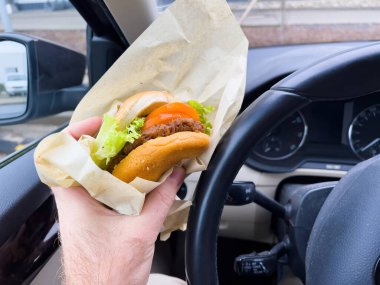 İçinde domates dilimi olan lezzetli bir hamburger tutan bir insan, arabasının içinde, hareket halinde olan bir ulaşım aracı için yapılmış fast food..