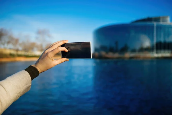 A woman captures the view of a building by the sea, holding a portable device. The defocused government building adds distinction to the scene.