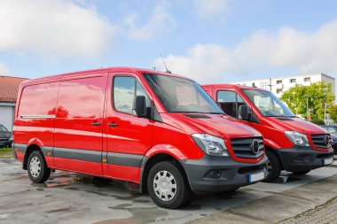 Şehir merkezine park etmiş iki kırmızı teslimat aracının yan görüntüsü mavi gökyüzü ve üzeri dağınık bulutlarla dolu. Lojistik ulaşım ürün ve servis konsepti.