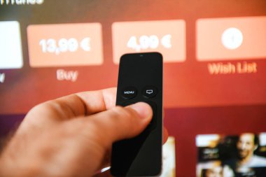 Paris, Fransa - 25 Kasım 2015: Bir TV ekranı önünde uzaktan kumanda tutan bir erkek eli ile kırmızı bir alçı sergiler. Arka planda, satın alma ve kiralama seçenekleriyle odaklanmış bir televizyon var.